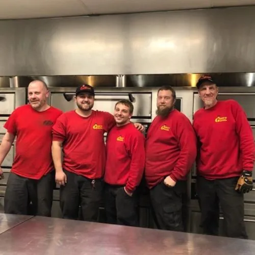 Five technicians in company shirts standing in front of a stainless oven line in a commercial kitchen